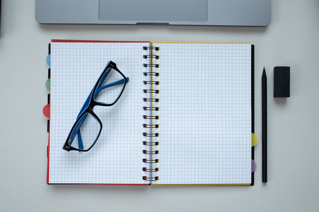 Top view of an open grid notebook, eyeglasses, and stationery arranged neatly on a desk.